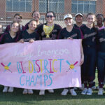 Girls' softball team posing with a trophy and a pink banner reading 'Lady Broncos Be District Champs' on a sunny field behind a chain-link fence.