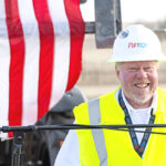Construction worker in a yellow safety vest and white hard hat speaks at a podium, with a large American flag in the background.