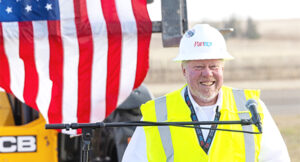 Construction worker in a yellow safety vest and white hard hat speaks at a podium, with a large American flag in the background.