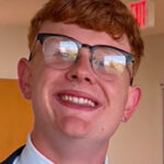 Portrait of a smiling young man with short red hair and glasses, wearing a dark suit, light blue shirt, and tie, indoors.