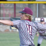 Baseball pitcher in a gray pinstripe uniform and maroon cap winds up to throw on the field, ball in his raised right hand.