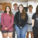 Nine students pose for a group photo in a classroom, wearing casual clothing and facing the camera.