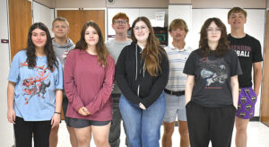 Nine students pose for a group photo in a classroom, wearing casual clothing and facing the camera.