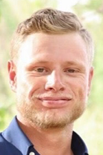 Close-up of a smiling man with short blond hair wearing a blue shirt, outdoors.