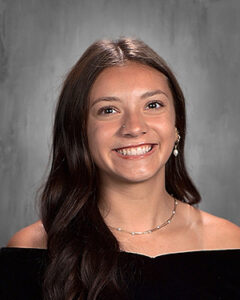 Portrait of a smiling young woman with long dark hair, wearing a black off-shoulder top, against a gray backdrop.