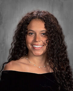 Smiling woman with long curly dark hair, wearing an off-the-shoulder black top and a delicate necklace, posed against a gray studio backdrop.