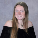 Portrait of a young woman with long light brown hair and blue eyes, wearing an off-the-shoulder black top and a necklace, smiling against a gray studio backdrop.
