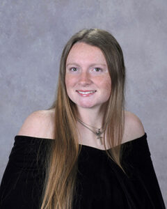 Portrait of a young woman with long light brown hair and blue eyes, wearing an off-the-shoulder black top and a necklace, smiling against a gray studio backdrop.