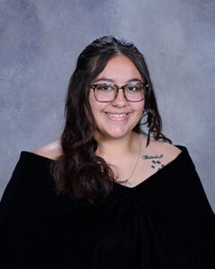 Smiling young woman with glasses and long dark hair, wearing a black off‑the‑shoulder top; left shoulder shows a paw‑print tattoo.