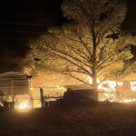 Night scene of a rural property on fire with flames around sheds and a large tree, smoke rising in the dark.