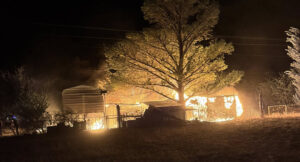 Night scene of a rural property on fire with flames around sheds and a large tree, smoke rising in the dark.