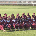 Outdoor graduation photo: graduates in maroon gowns and caps seated on a grassy field with bleachers in the background, some faculty standing at the edge.
