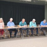 Eight adults seated at a long table on a stage, signing papers during a community meeting; a Texas flag is in the background.