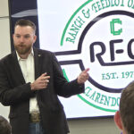 Male presenter in a dark blazer speaks to an audience, gesturing with his hands as a large screen displays a circular green-and-black logo behind him.